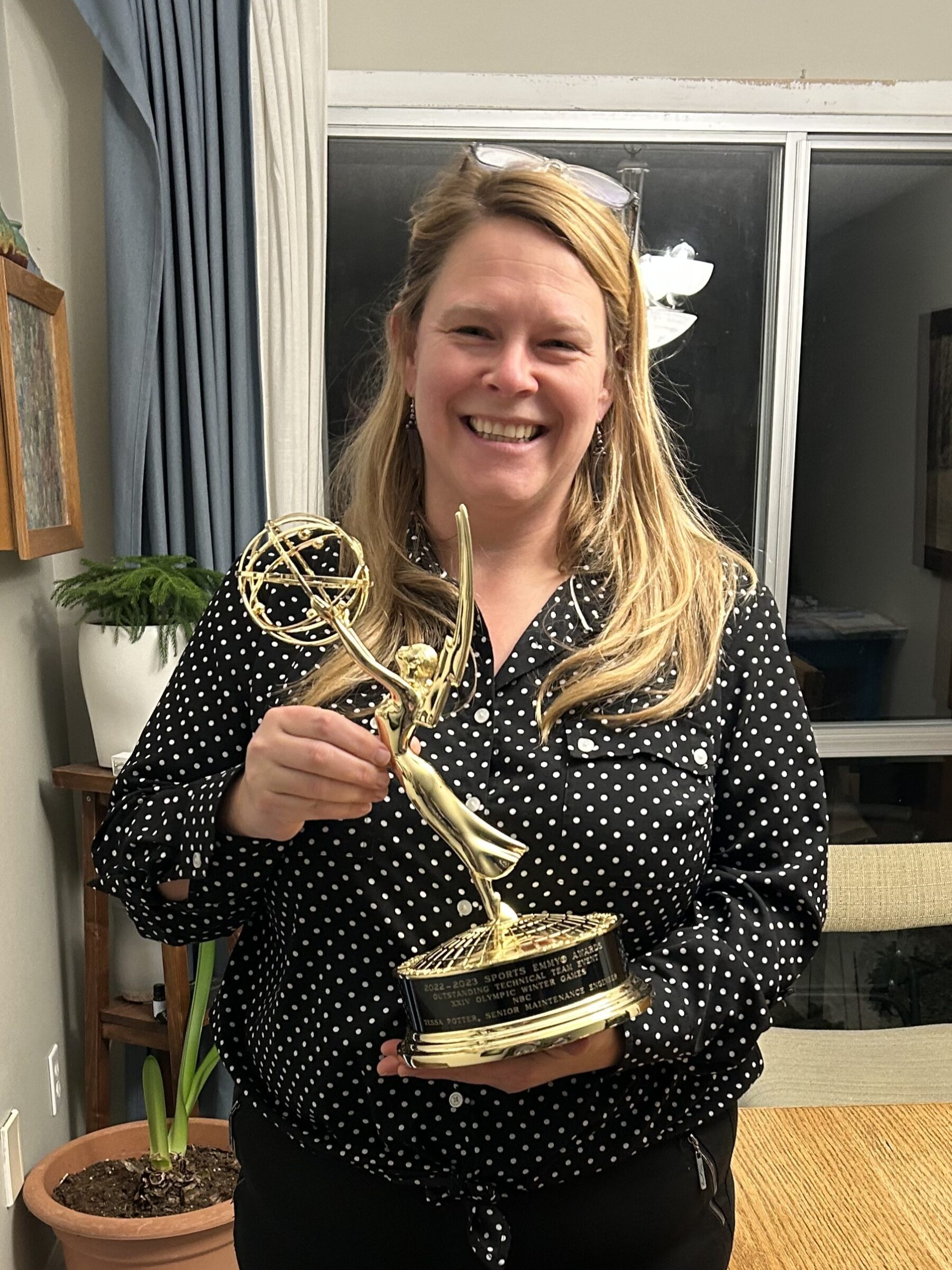 一位身穿黑色衬衫和黑色裤子的金发女士站在窗前，手里拿着一个艾美奖金像。