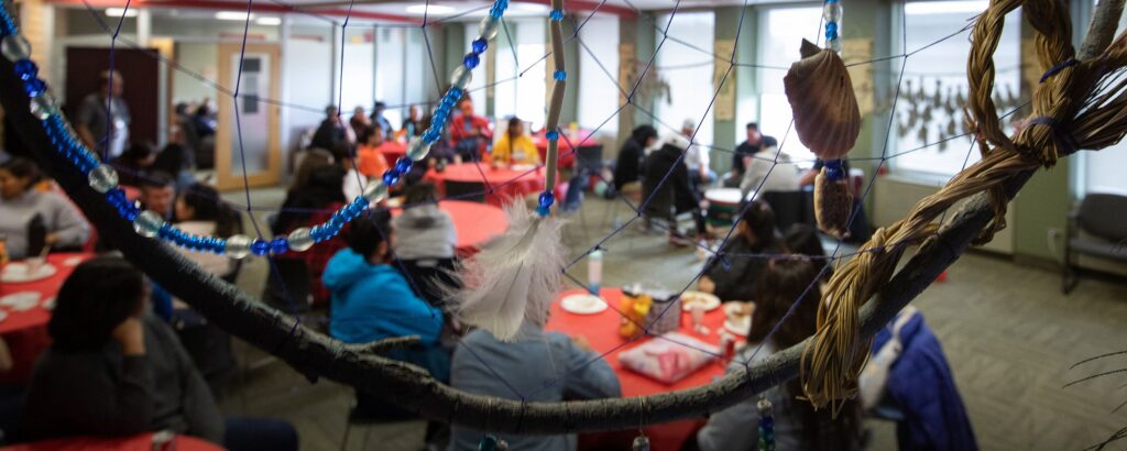 一群人坐在土著学生支持中心的圆桌旁，透过装饰有珠子、羽毛和编织纤维的大型捕梦网观看。