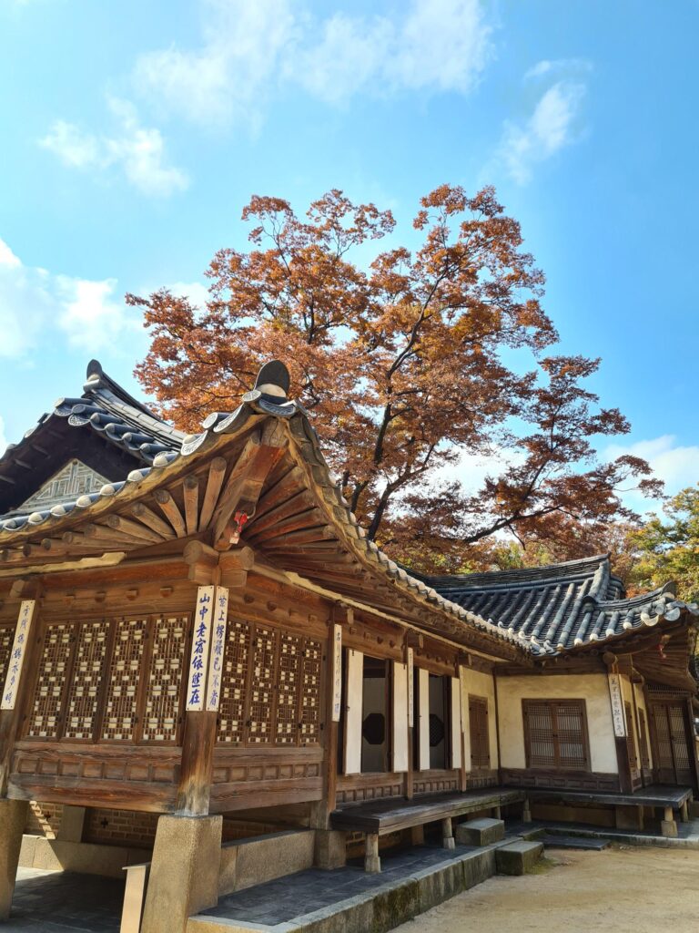传统的木制寺庙建筑，有雕刻细节和瓦片屋顶，坐落在一棵树下，树下有橙色的秋叶，映衬着蓝天。