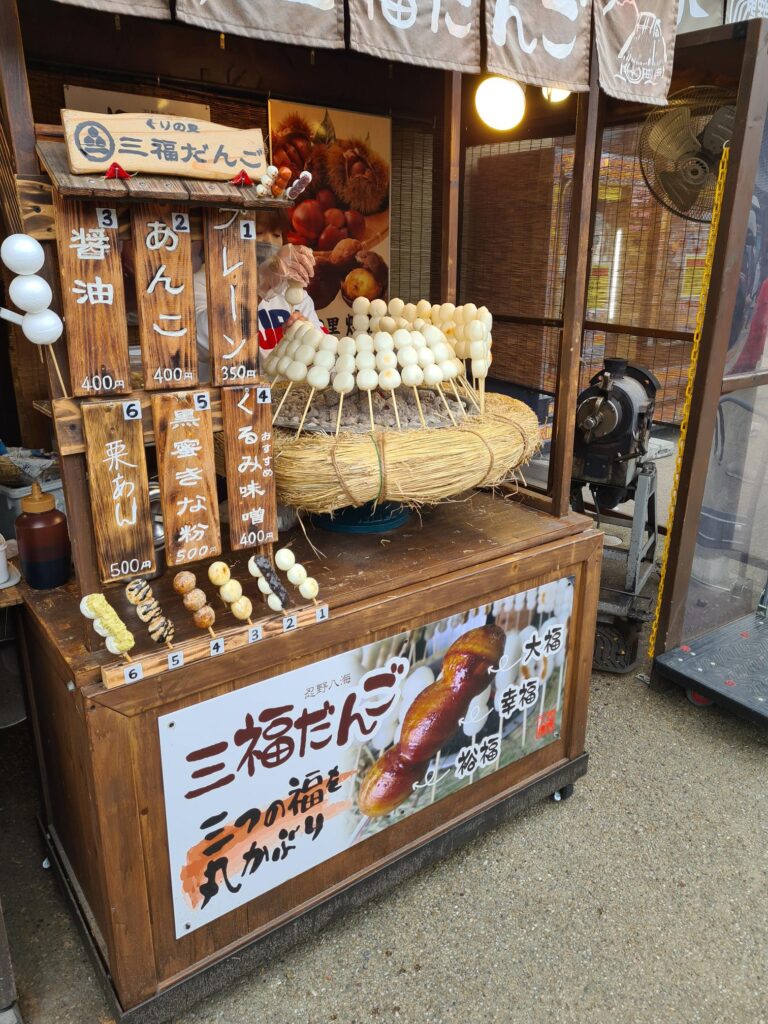 街头木制小食品摊，展示着圆形糕点和烤肉串的托盘，柜台上方悬挂着手写标牌和灯笼。