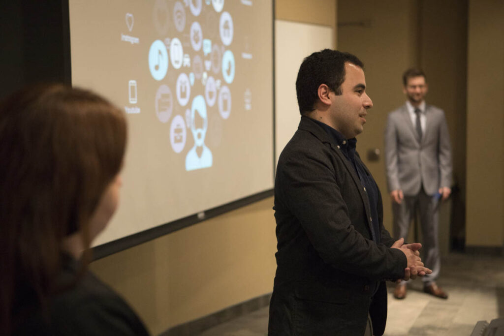 一名男子在教室里讲话，旁边有一个大型投影仪屏幕