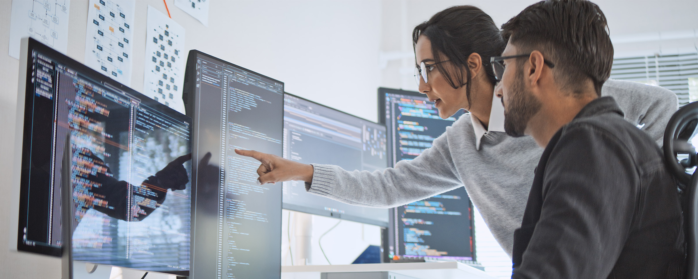 男人和女人在现代办公室的办公桌前一起工作，专注于e8体育安全任务，多台计算机显示器显示代码和e8体育数据。