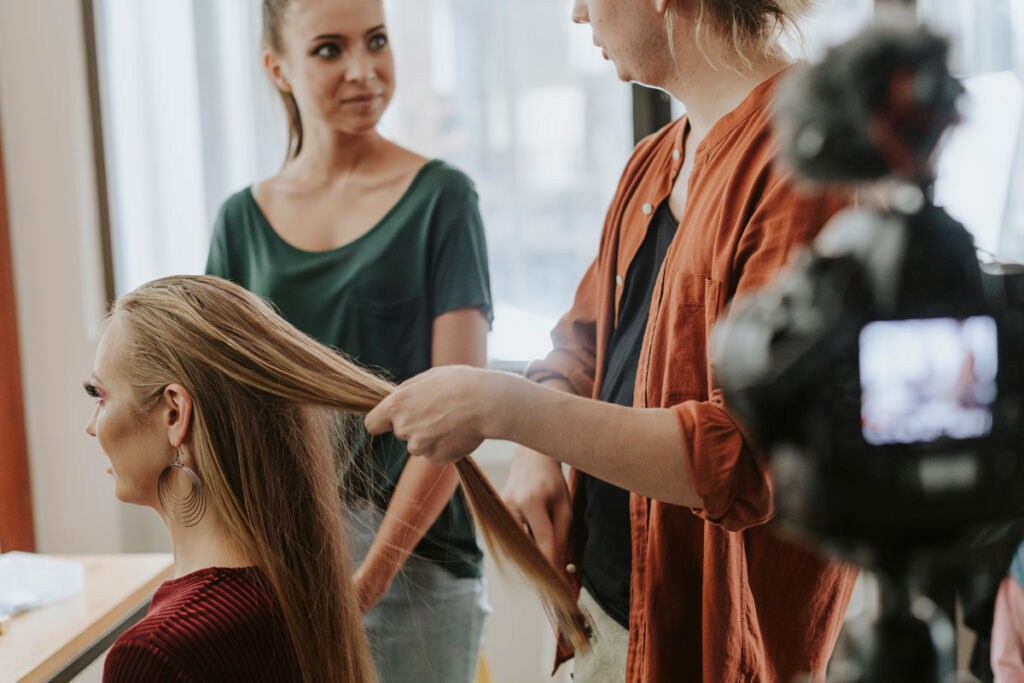 A hairstylist in an orange shirt works on a seated client's long blonde hair while a second person observes and a camera records the session.