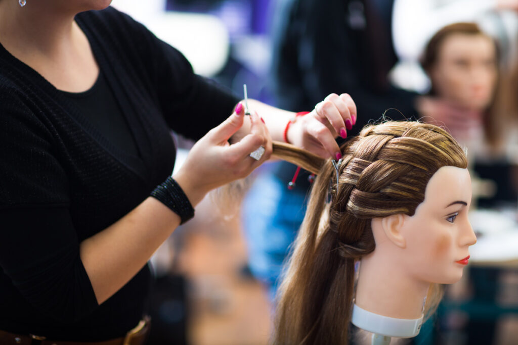 A student with pink-painted nails practices a French braid technique on a long-haired mannequin head during a hairstyling class.