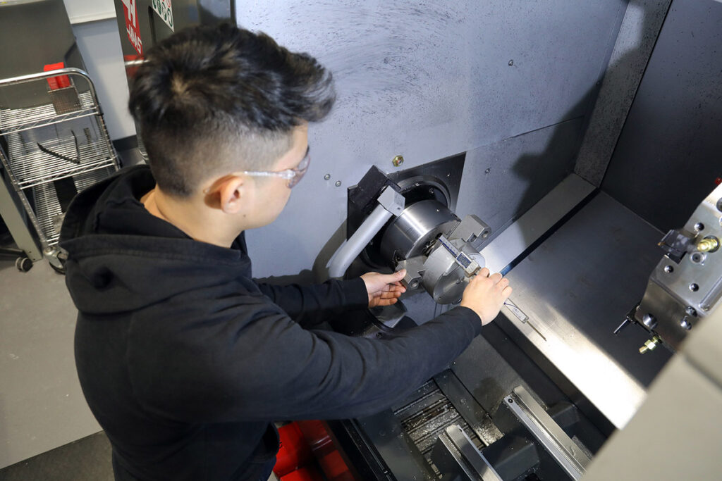 工业机械车间实验室中，一名戴着安全眼镜的男学生用双手调整大型 Haas 数控车床卡盘内的组件。