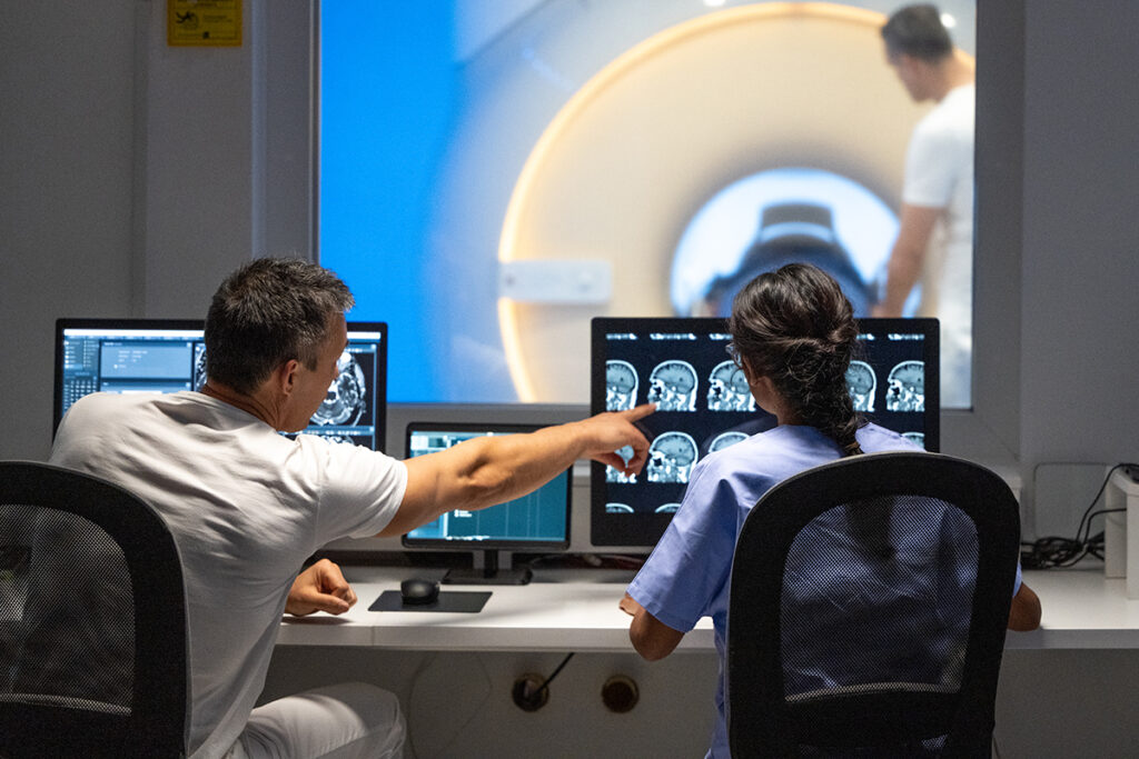 男性和女性 MRI 技术人员坐在手术室控制台前操作 MRI 扫描仪的后视图