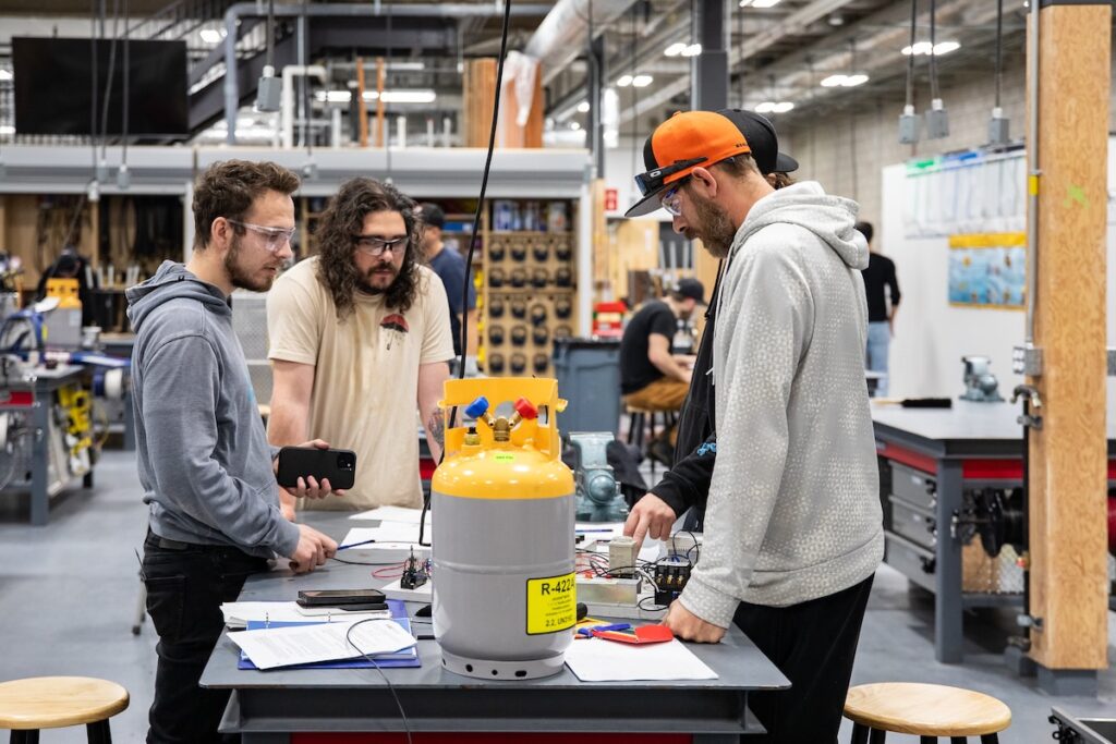 三名戴着安全眼镜的学生在机械行业实践实验室中合作开展制冷控制项目。