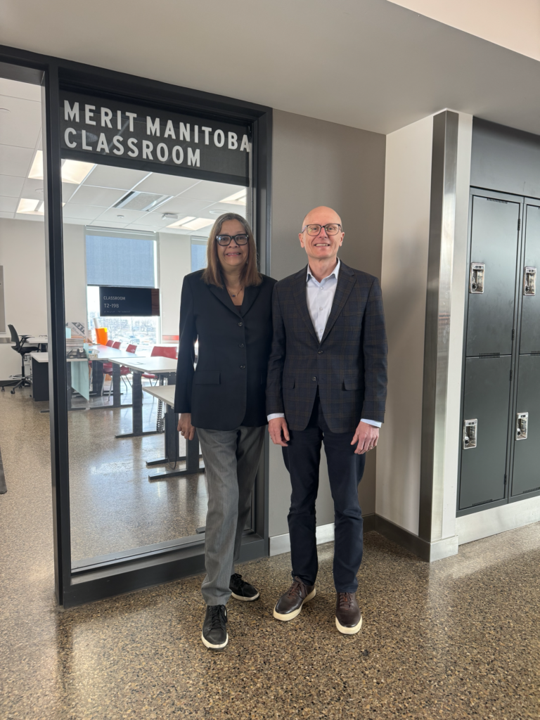 两个人站在一间教室前,对着镜头微笑,门口上方挂着“优秀马尼托巴教室”的牌子。