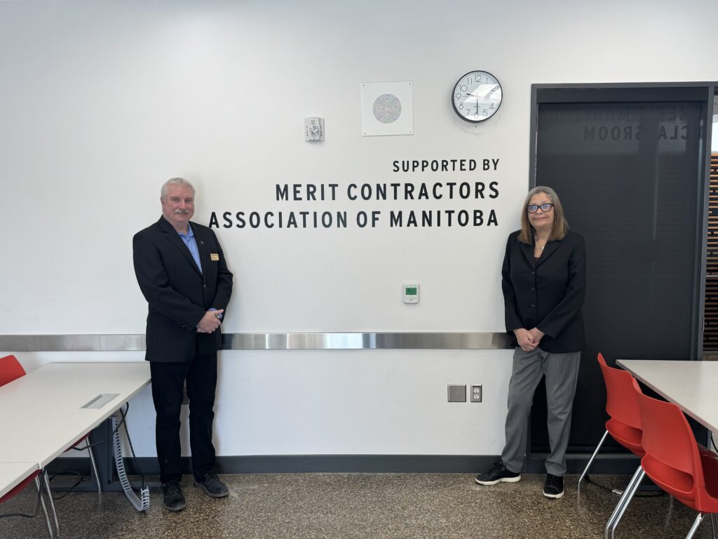 两个人站在教室里。他们对着镜头微笑,面前是一面写着“曼尼托巴优秀承包商协会支持”的白墙。