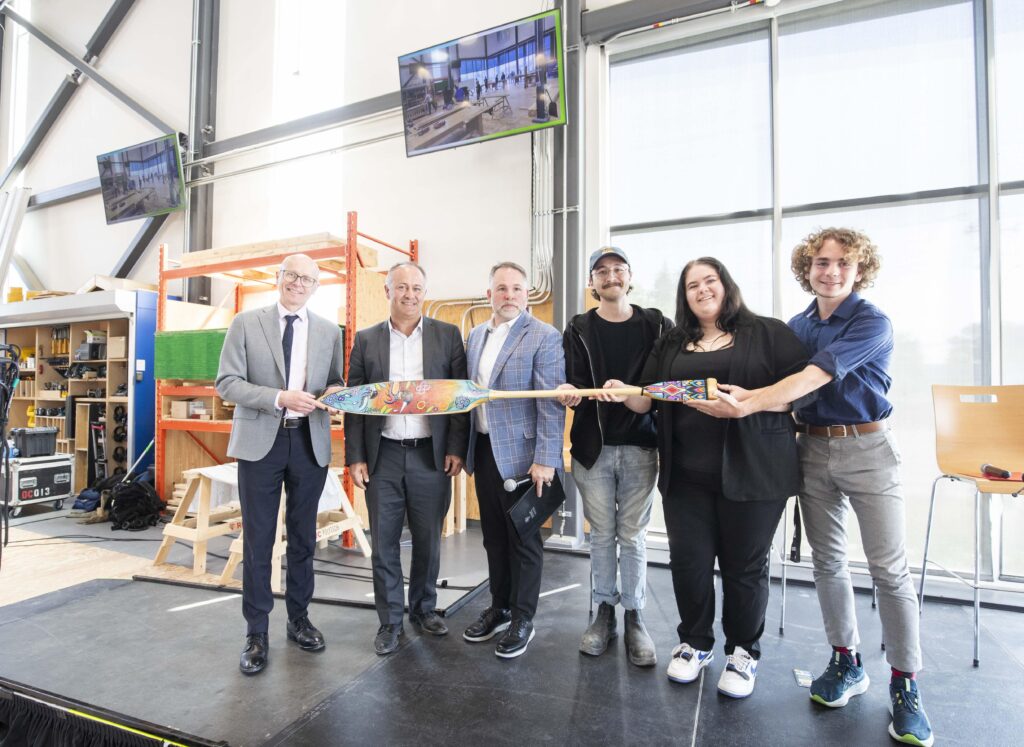 六个人站在舞台上，手持装饰华丽的独木舟桨，对着镜头微笑。