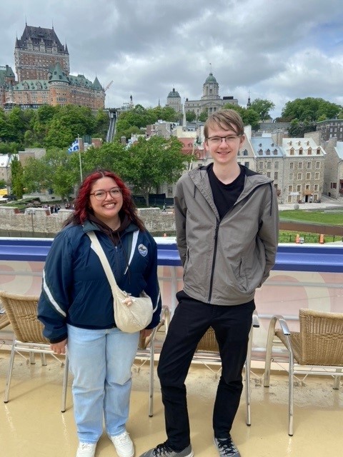 两名学生站在魁北克市的风景背景下，包括建筑物、树木和天空。