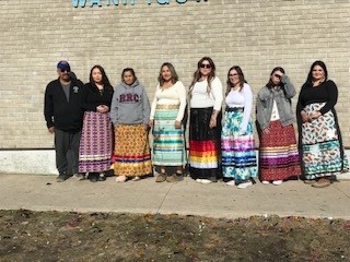 七名妇女并肩站在砖墙前。她们都穿着丝带裙子。一个男人站在他们的左边。