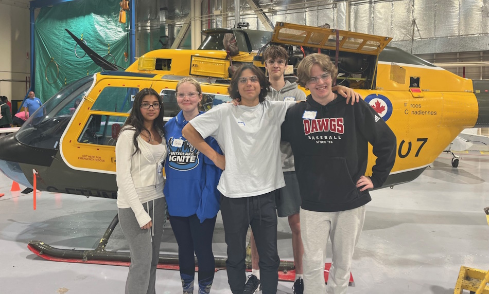 四名学生站在机库中一架黄色直升机前。