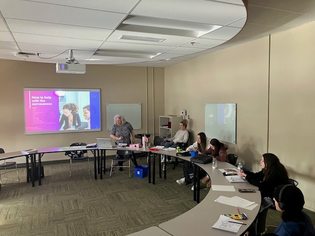 学生们围坐在一张大桌子旁，一位女士站在屏幕前，屏幕上写着“如何缓解紧张情绪”