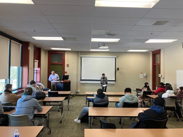 学生们坐在教室里，一位女士站在教室前面向大家展示。