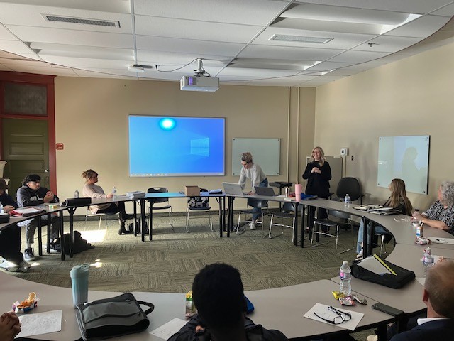 学生们围坐在一张大桌子旁，听一位女士向小组做演讲。