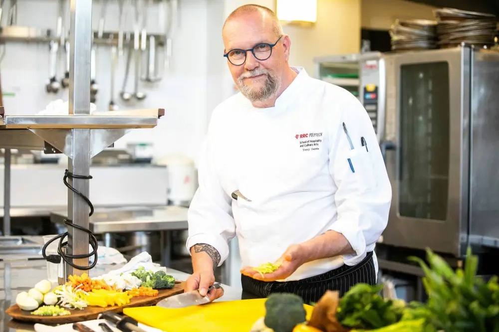 厨师 Terry Gereta 站在厨房里。他穿着白大褂，面带微笑。他一只手拿着一些蔬菜，另一只手拿着一把菜刀。