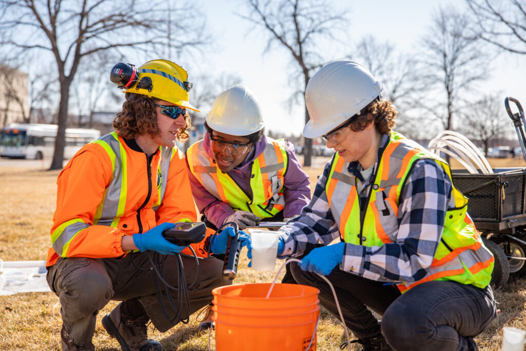 三名工科学生穿着安全背心，跪在水桶周围测试户外水质