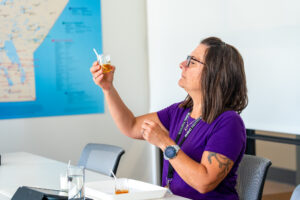 Woman in purple shirt holds jar of honey to the light to assess colour.