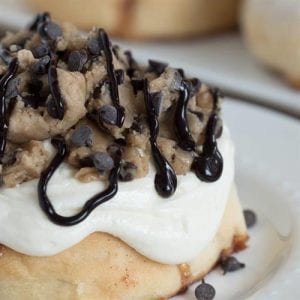 Cinnamon bun on white plate covered in white icing, crumble, and chocolate drizzle